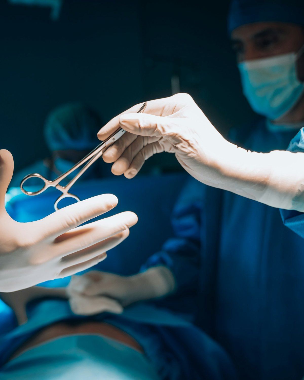 A gloved hand passing a surgical hemostat to another gloved hand across an operating theatre, a masked surgeon looking on