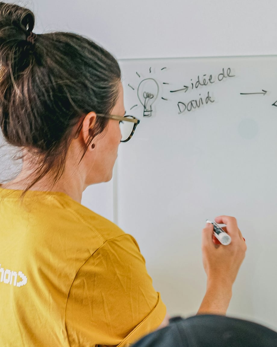 A woman in a yellow t-shirt drawing a flow of boxes and arrows on a whiteboard, a colleague watching in the foreground