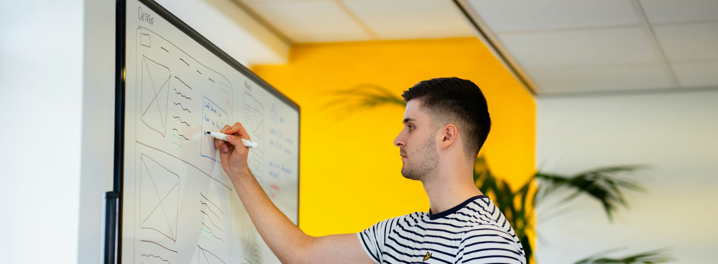 A man sketching screen layouts on a whiteboard against a yellow wall