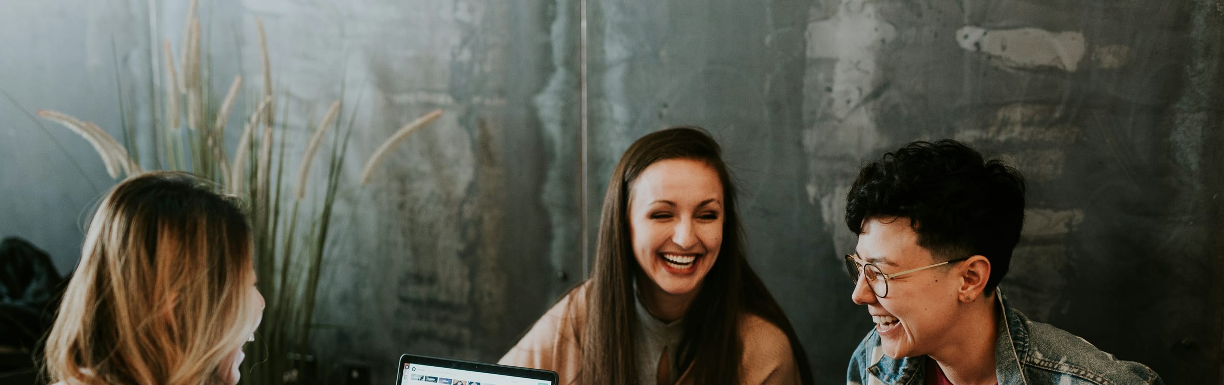 Three colleagues laughing over their laptops at a table against a bare concrete wall