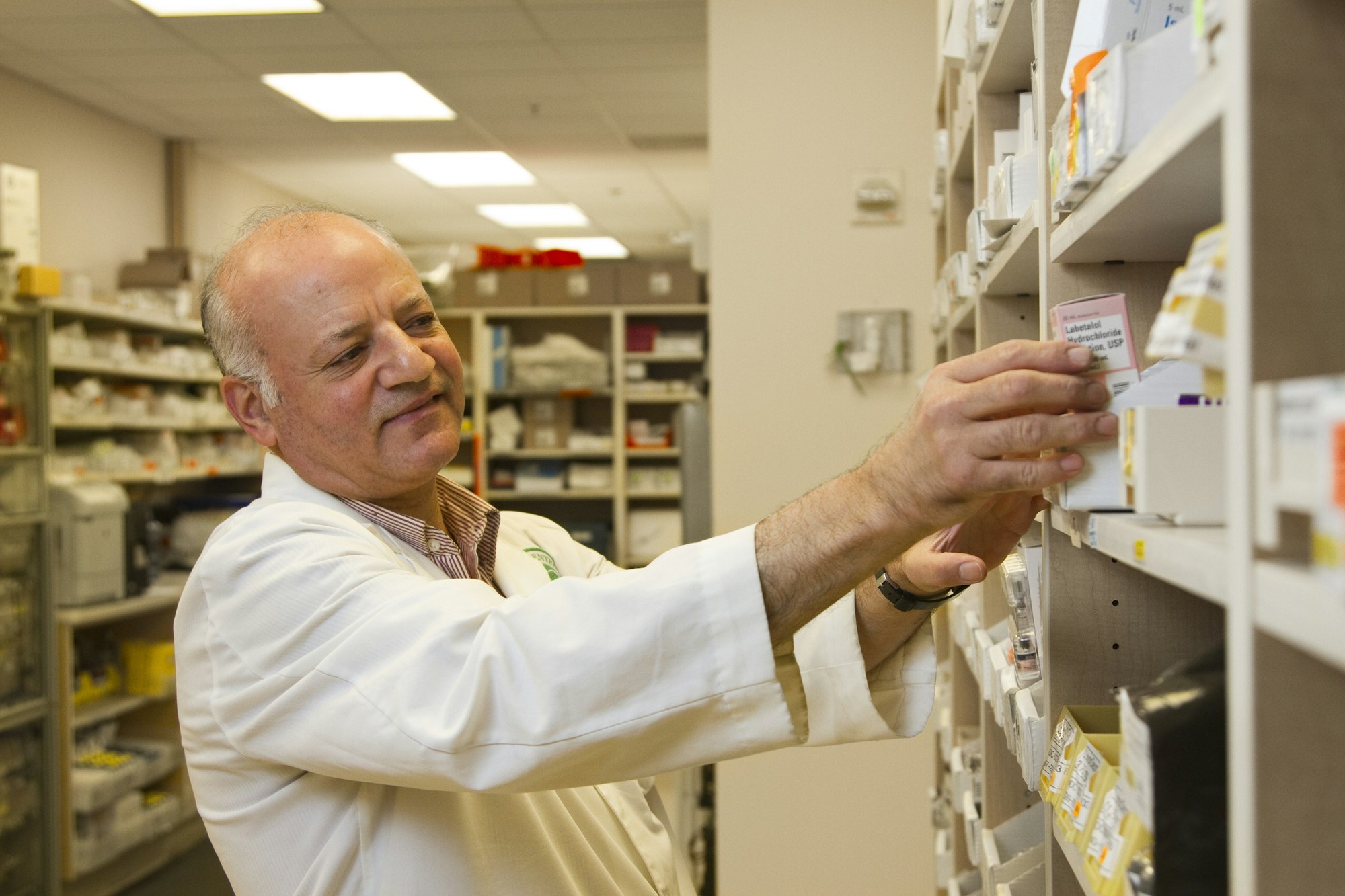 A pharmacist in a white coat smiling as he takes a medicine carton from a dispensary shelf