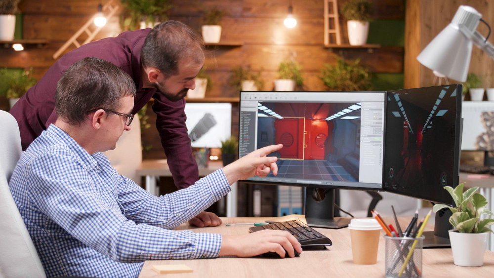 Two engineers at a desk, one pointing at a 3D scene on a monitor while the other leans in